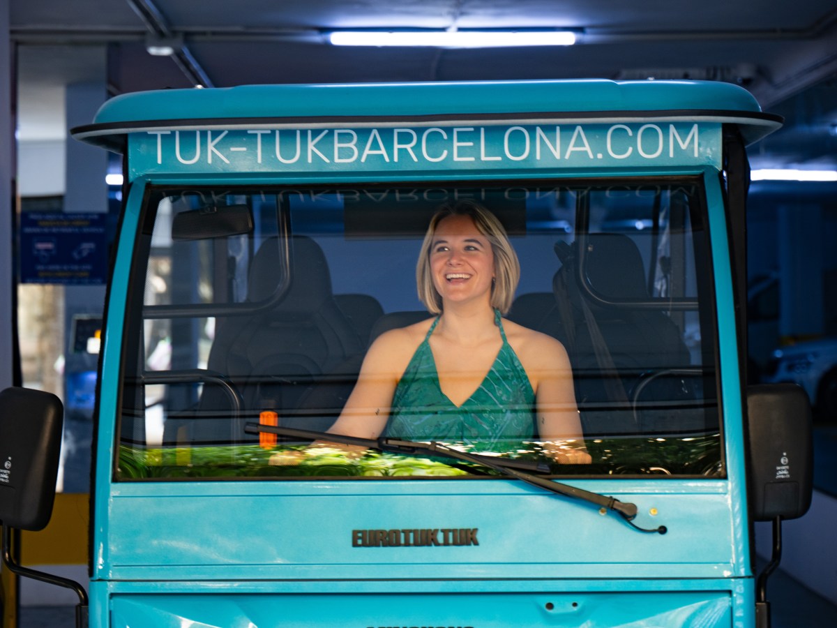 Smiling person in a turquoise tuk-tuk with 'TUK-TUKBARCELONA.COM' on the roof in a parking garage.