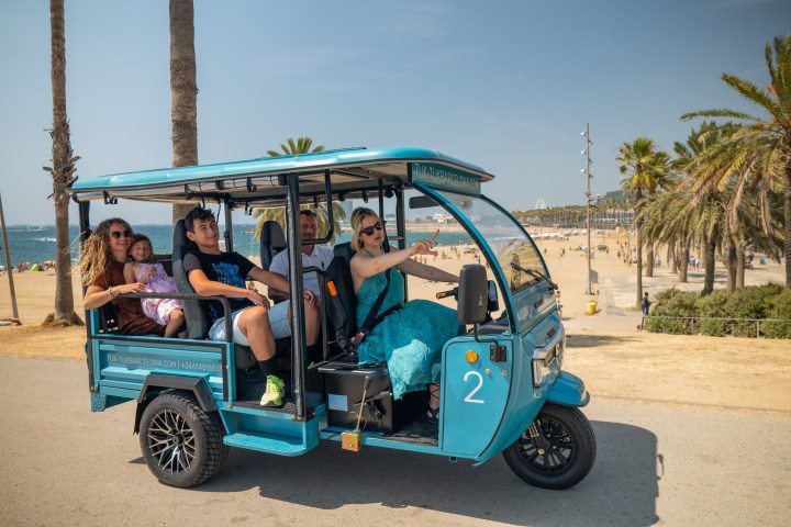 People riding a blue tuk-tuk near a beach with palm trees and clear skies.
