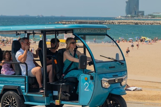 Blue tuk-tuk on a beach road with people inside, beach and sea in the background.