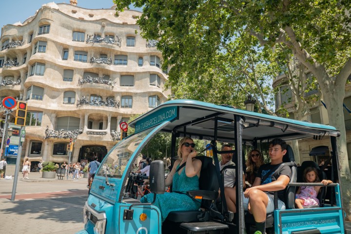 People on a blue tuk-tuk in front of a modernist building with wavy façade.