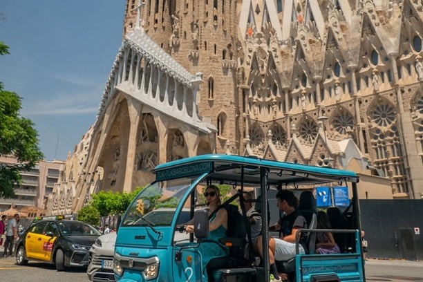 Blue tuk-tuk with passengers in front of the Sagrada Familia in Barcelona on a sunny day.