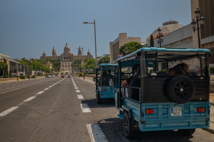 Street view of blue electric tuk-tuks driving towards a grand building in Barcelona.