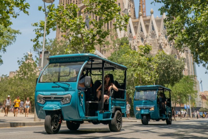 Two blue tuk-tuks drive on a sunny tree-lined street with a cathedral in the background.