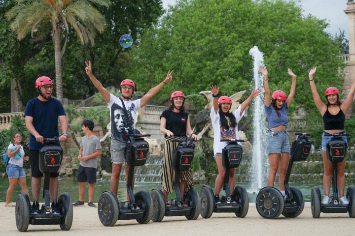 Five people in pink helmets on Segways, posing joyfully near a fountain and trees.