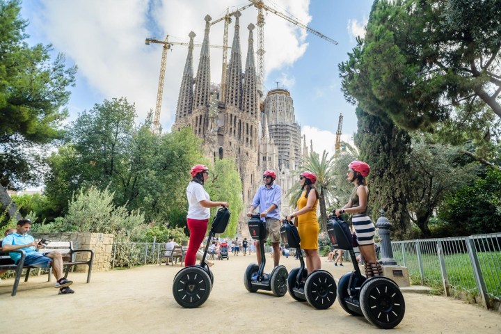 Four people on segways near a cathedral under construction, with cranes and trees around.